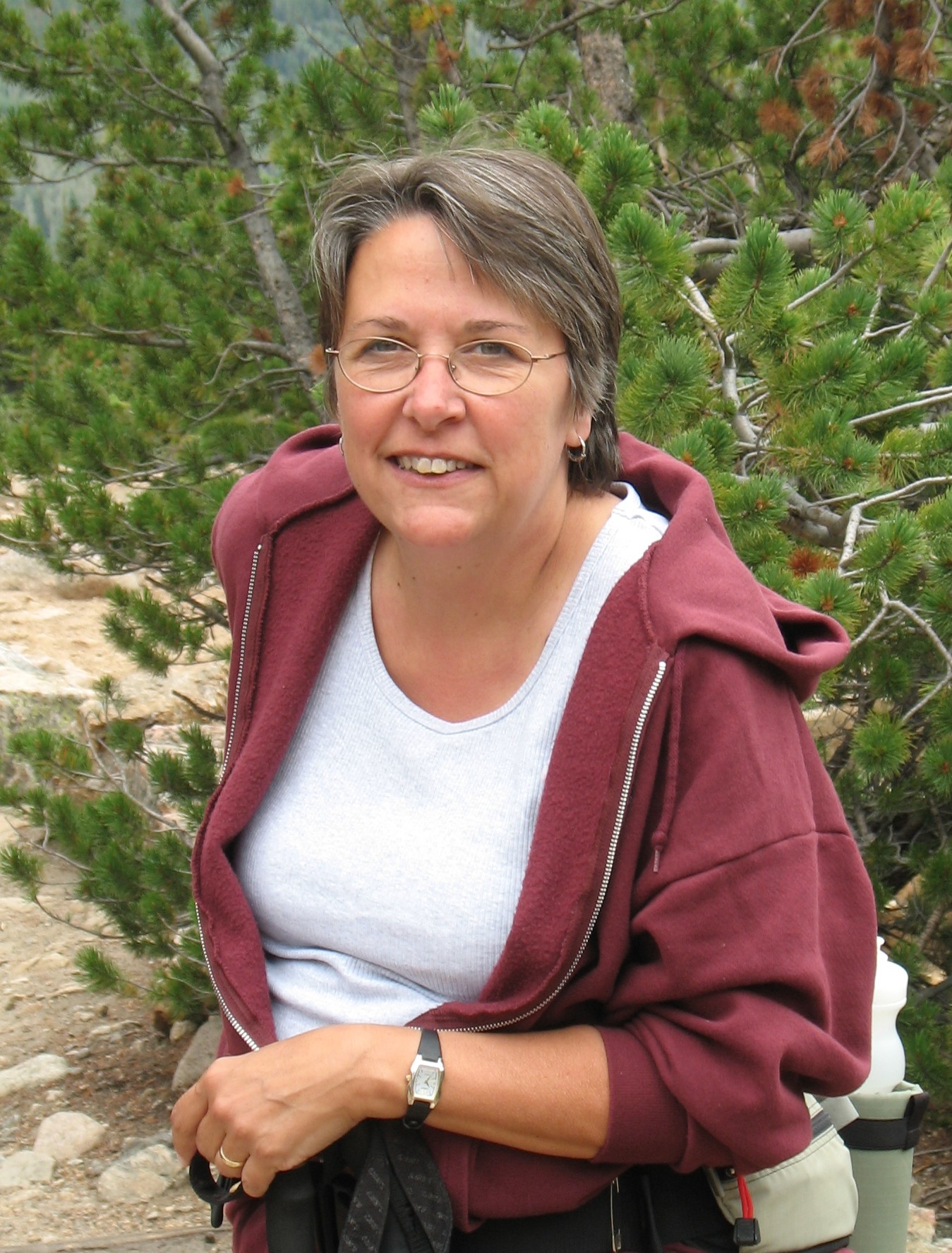 A person with short hair, glasses, and a maroon hoodie is smiling outdoors. Pine trees and rocky terrain are in the background.